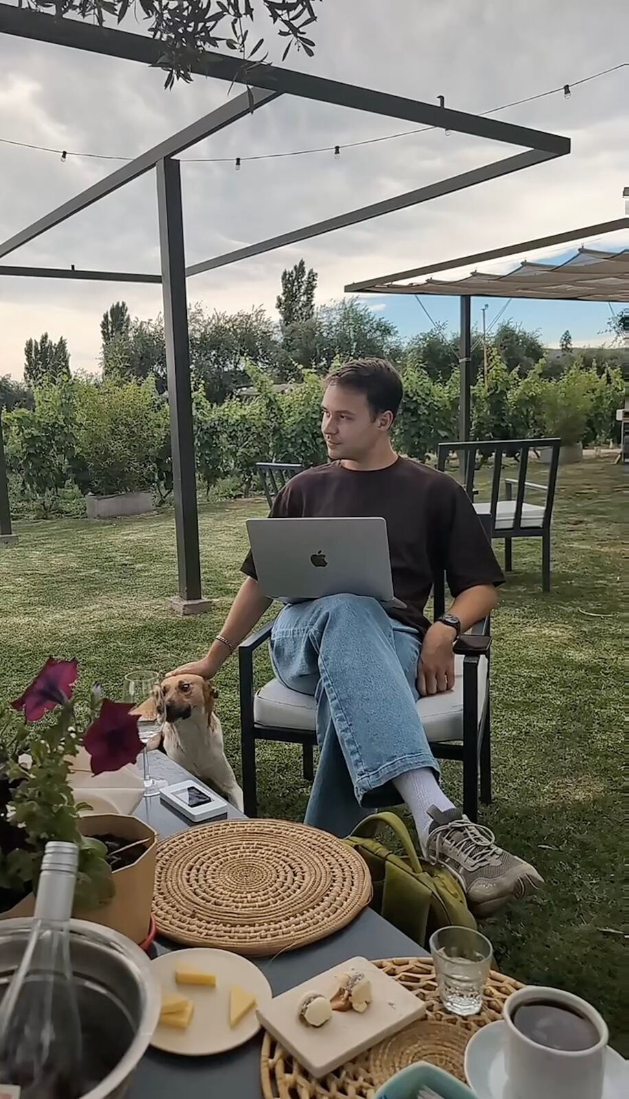 Elijah working on a laptop at a vineyard, petting a dog